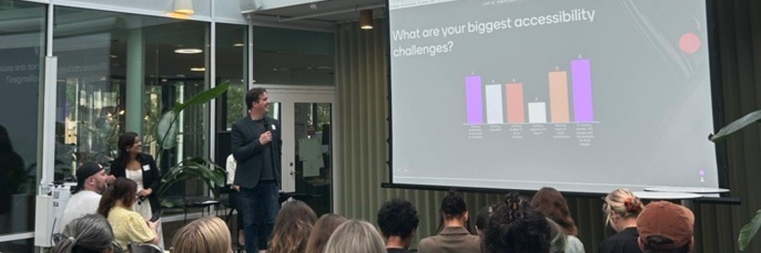 Chris stands in front of an audience of seated people, holding a microphone. On a screen is a slide reading 'What are your biggest accessibility challenges?', and the results of a Menti survey.